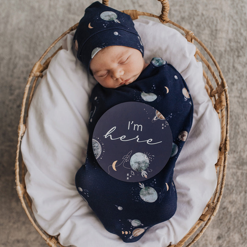 The Milky Way Snuggle Swaddle in navy blue with the milky way & planets design. Matching beanie included