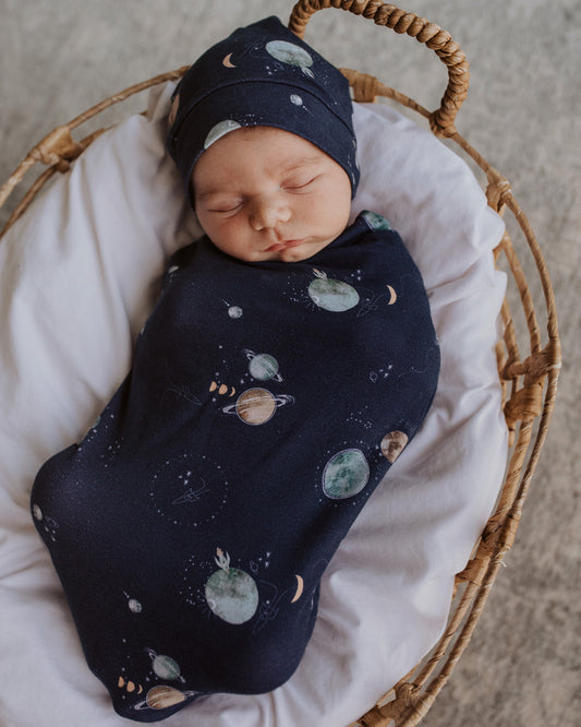 The Milky Way Snuggle Swaddle in navy blue with the milky way & planets design. Matching beanie included