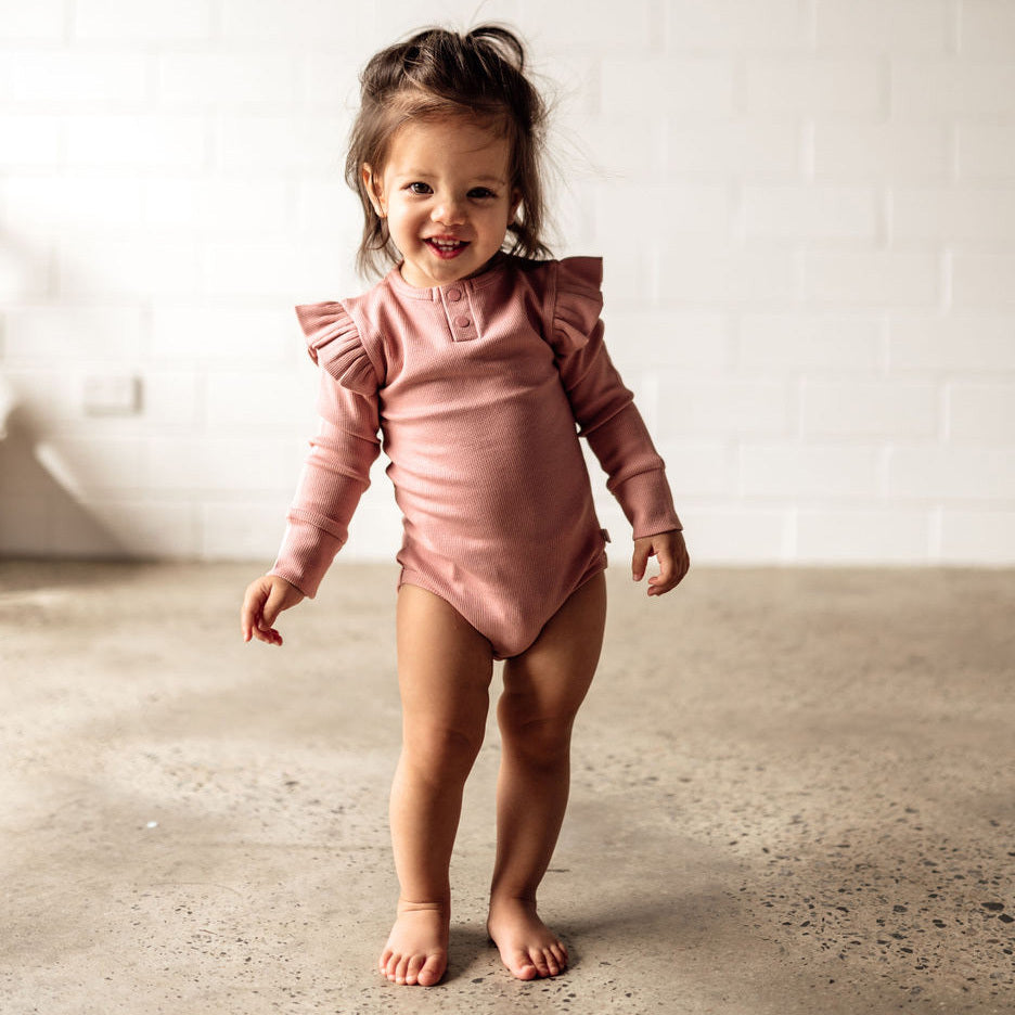 Toddler in the dusty rose pink body suit in organic rib fabric, It has small frills on the shoulders & 3 button clips in the crutch for easy nappy changes.. has fold over cuffs to keep hands warm